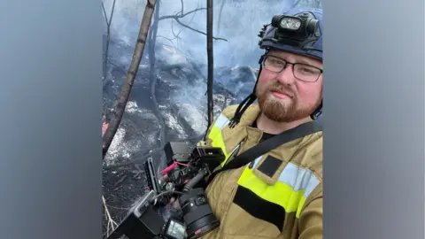 Ed Matthews, who wears a protective uniform, holds a camera. He is stood in a smoky woodland area.