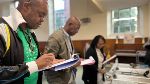 Jon Wright/BBC Judges Brian Powlett, Frank Clarke and Ivya Scott stand in front of a row of food dishes with clip boards