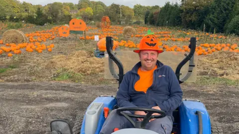 A man in an orange t-shirt, pumpkin decorated hat, blue fleece and blue trousers sits on a tractor. Behind him is a field filled with pumpkins, a couple of haybales and two large Jack-o'-Lanterns made from wood and paint. The field is surrounded by trees.