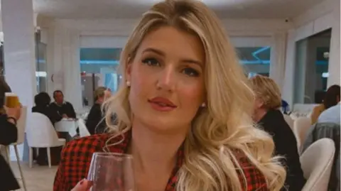 A woman with long blonde hair looking into the camera, while seated at a restaurant table. She has a black and red check jacket on and is holding a wine glass in her right hand. 