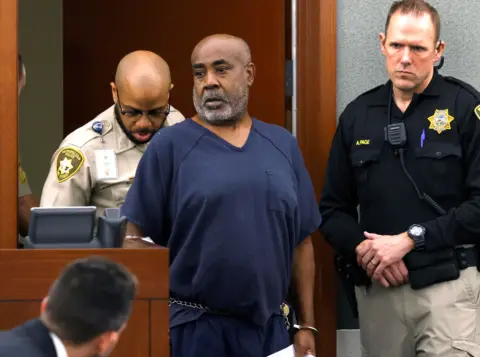 Reuters Duane Davis appears in court. He is wearing a loose-fitting blue t-shirt, and his hands are cuffed with a chain passing in front of his waist. Police officers flank him at either shoulder
