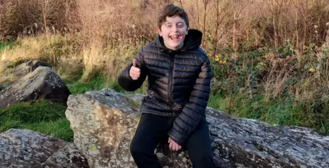 13-year-old Kyle sitting on a rock overlooking his hometown, Pontypridd. He has short black hair and is wearing a black puffer coat. He is smiling and making a thumbs up gesture.