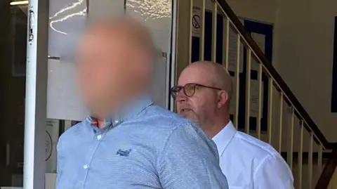 George King/BBC Andrew Wickenden wearing glasses and a white shirt leaving court behind another man who is wearing a blue shirt. The face of the second man, in the foreground, is blurred out.