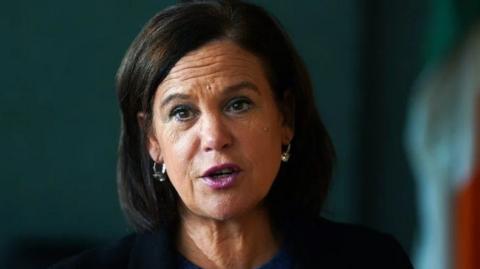 Mary Lou McDonald mid conversation with pearl drop earrings on and a blurred irish flag in the right hand corner 