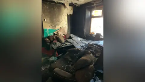 Paul Fosh Auctions The inside of the same terraced house in another room, with new debris including a burnt and dirt covered sofa and peeling burnt wallpaper 