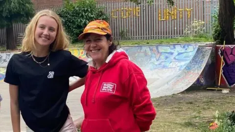A young woman with shoulder-length blond hair, wearing a black t-shirt branded with "SB GB" stands smiling next to an older woman wearing a brightly coloured baseball cap and a red hoodie. Both are smiling at something behind the camera and are standing in front of a skateboard ramp which has been decorated with graffiti.