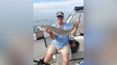 David Payne pictured holding a fish. David is sat on a fishing boat out at sea.
