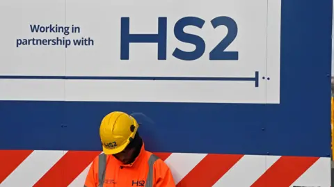 Reuters An HS2 workers in orange hi-vis clothes and a yellow hard-hat sits in front of blue, red and white hoarding with an HS2 sign on it