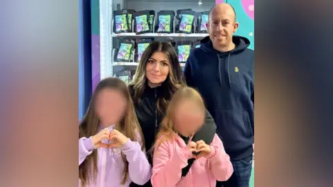 A family photo of Amy and Derek and their two daughters standing in front of one of their vending machines. The girls are wearing light purple and pink hoodies and doing 'heart' hand gestures