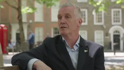 A man in a suit and light blue shirt sits on park bench talking to a reporter. A row of Regency-style terraced houses can been seen in the background. 