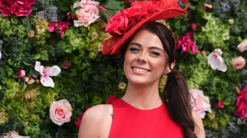 PA Media A racegoer stand in front of a wall of flowers. She wears a red dress and red fascinator hat.
