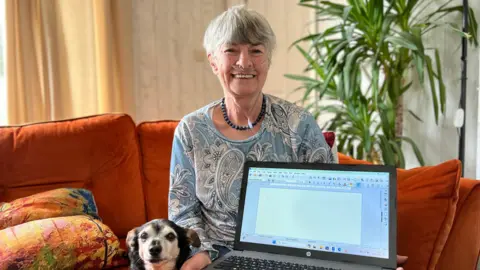A woman sits on her orange sofa with a laptop on her lap. She is smiling and her dog is sitting beside her. There is a large plant behind her. She is wearing a long-sleeved blue and white paisley top and has short grey hair.