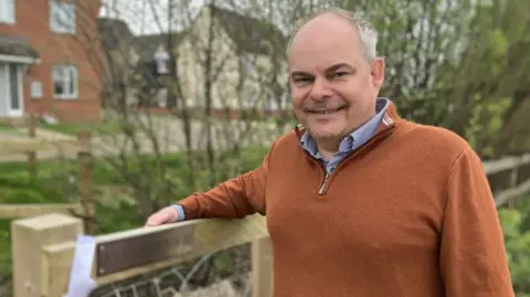 Councillor Ian Snowdon smiling for the camera in front of a residential area. He rests his right arm on a fence.