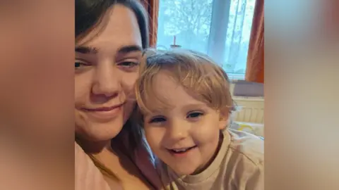 Abi A mother takes a selfie with her young son. He is leaning his head into her cheek and they are both smiling. He has blonde hair and is wearing a cream T-shirt. She has dark brown hair. They are inside a house with a window and curtains in the background.