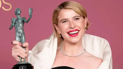 Buckley has short blonde hair and pink lipstick on. She is wearing a black and white dress and is holding up her grey award.