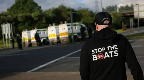 Reuters A man wears a 'Stop the Boats' hoody, as police and their vehicles block a road to stop anti-immigrant protesters from reaching Sandyknowes Roundabout.