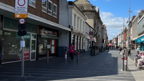 A view of Grimsby town centre shows rows of shops on either side under a blue sky. On the left, an empty store with a red "To Let" sign in the window.