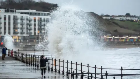 PA Media Woman standing on quayside photographing large wave coming towards her