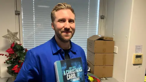 James Grant/BBC A man in a blue shirt holds up a script of A Christmas Carol. Behind him is a small Christmas tree and three cardboard boxes piled on top of each other.