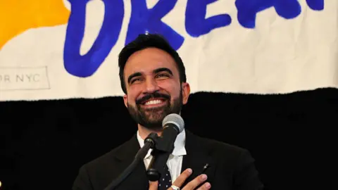 New York mayoral candidate, State Rep. Zohran Mamdani in front of a mic with hand on his heart.