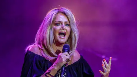 Bonnie Tyler on stage in purple pink lighting wearing black top and holding microphone. She has long blonde hair worn down.