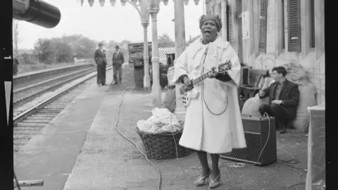 Sister Rosetta Tharpe stands on th epaltform in a large coat playing an electric guitar on the station platform.