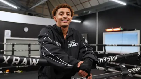 Saad Abdullah smiling towards the camera in a boxing ring. He is wearing a black jacket that has Team Saad on it and is in front of a black-themed boxing gym. 