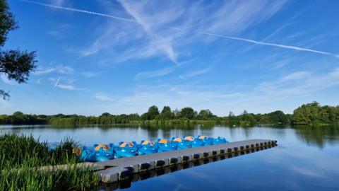 WEDNESDAY - A lake with a line of blue boats along a pontoon at Woodley. There are green plants in the water on the left of the frame and on the far side of the lake there are trees with green leaves against the blue sky.
