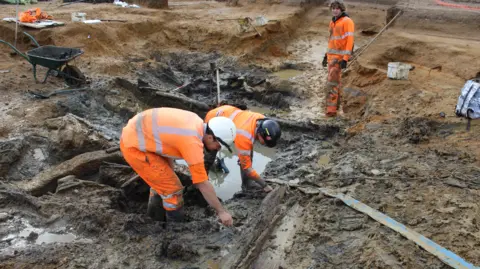 Cotswold Archaeology 3 archaeologists in head-to-toe orange high visibility clothing and hard hats searching through a muddy pit to deeper uncover the 3500-year-old remains of a wooden watering hole.