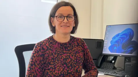 BBC/ Alex Osborne Image of Dr Lucy Joslin - pictured in her clinic beside a desk and computer, wearing an orange and pink spotted jumper, with glasses and dark short hair.