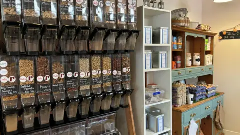 The inside of Roots cafe. The wall is covered with refill containers of nuts, beans, seeds and cereals. The is a white shelving unit in the middle with metal tins of flour and other dry ingredients, and on the right there is a blue dresser with stacks of egg cartons. 