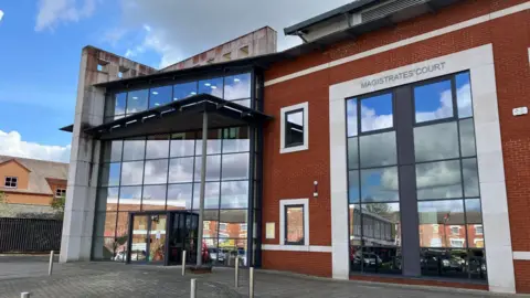 A large brick and glass building on a sunny day. The left half of the building is made of glass panels and has a revolving door on the ground floor. The right half is made of red bricks and glass panels, with a stone sign saying "MAGISTRATES' COURT" in capital letters.