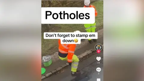 Unknown Two men in orange and yellow hi-vis with white helmets are at the side of a road on which a pothole has been filled. One is watching from the pavement while the other appears to be jumping on the pothole. Both feet are off the ground and his knees are bent.