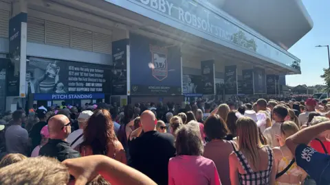 Alice Cunningham/BBC A large crowd of people wait outside Portman Road stadium. 