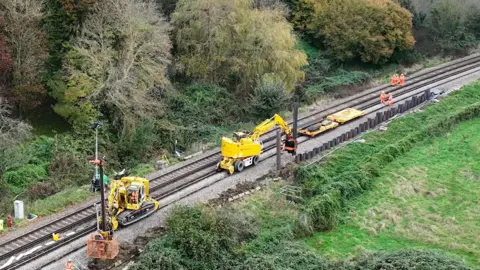 NetworkRail Yellow diggers are on a railway track where on one side a wall made of metal planks is being constructed. There are workers in hi-viz on the tracks as well and trees seen on the far side of the track