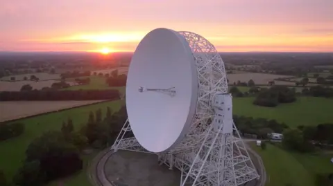 An aerial view at sunrise of the Lovell telescope at Jodrell Bank. Its huge white dish, taller than a house, is tilted upright on a steel cradle that sits on a circular track. Behind it, flat Cheshire farmland stretches to the horizon under a soft pink sky.


