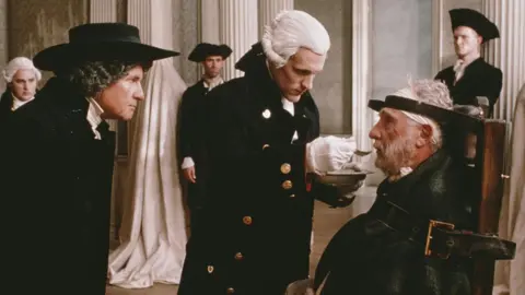Getty Images A film still showing men wearing late-18th Century clothes in a large room resembling a palace. On the left a man wears a black hat and jacket. He is staring at a man sitting in a chair wrapped in leather straps and a black straight-jacket. The seated man is being fed by a third man who wears a white wig and black jacket. Soldiers and servants stand in the background.