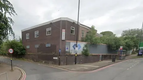 A Google street view showing a two-storey building with boarded up windows on the first floor. A sign on the corner of the building says Ladywood Social Club. Hoardings have been put up by a developer. The building is on the corner of a main road and side road, with street signs seen in the distance. A bus stop and lampposts can also be seen. It is daytime in the picture.