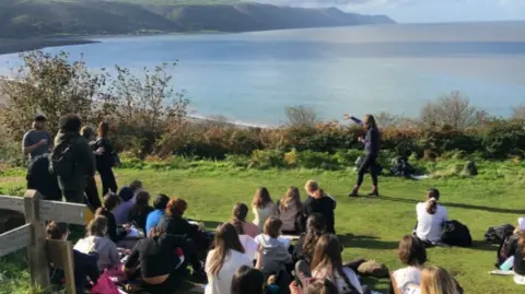 Field Studies Council Image shows a group of young people sitting on a green verge that looks over Porlock Bay in Devon. Some of the teenagers are standing, others are sitting. A person is stood in front of the group and pointing out to sea, as if they are telling the group some information. 