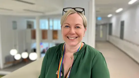 Laura Skaife-Knight standing in a corridor, wearing a green shirt. She has a rainbow lanyard round her neck. She's smiling and has blonde hair, with her glasses on her head. 