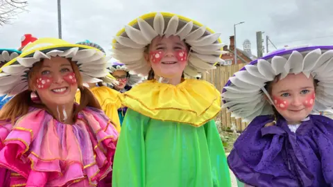 A woman and two young girls dressed in colourful costumes ahead of the St Patrick's Day parade in Belfast. They are wearing matching hats and their cheeks are painted red with white spots. 