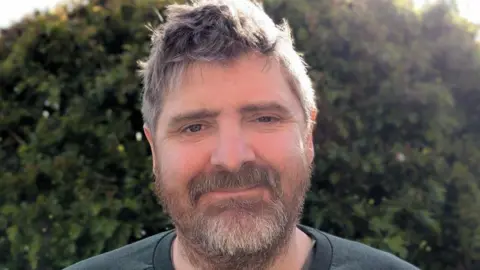 Ben Saffell Close-up portrait of Ben Saffell standing outdoors in front of a dense green hedge. He has short, slightly tousled hair and facial hair, and is wearing a dark crew-neck top. Bright sunlight from behind creates a soft glow around the head and shoulders.