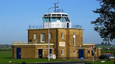 Glyn Baker/Geograph The control tower at North Weald Airfield, which is a brown brick building with a white tower on top. The tower offers panoramic views of the airfield.