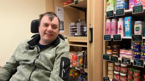 A man sitting in a wheelchair with brown hair and a green fleece. In the background there are shampoos and tins on shelves.