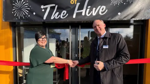 A woman with short black hair, wearing a green dress and black glasses, shakes the hand of a man in a black coat. They both smile, standing in front of a red ribbon which is tied across two glass doors. A black banner with fireworks on it hangs over them, saying 'The Hive'