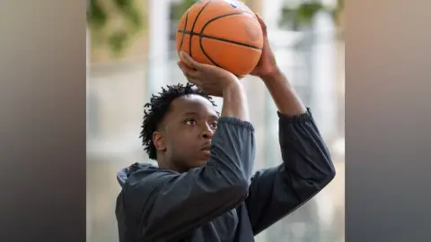 Se puede ver a GLA OG Anunoby sosteniendo un balón de baloncesto por encima de la cabeza con ambas manos, preparándose para lanzar en una cancha al aire libre.