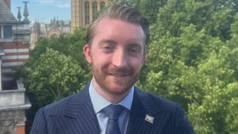 A man with a beard is wearing a mainly blue jacket, shirt and blue tie. Trees are in the background.