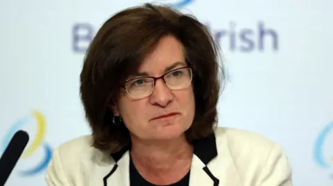 A woman with medium length brown hair and glasses wearing a white suit, Eluned Morgan