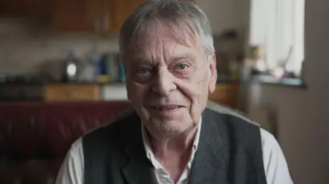 BBC An image of Ian Horne, aged 79, sat at home on the sofa, looking into camera and wearing a white shirt and grey waistcoat.