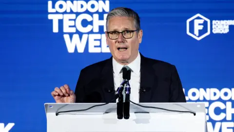 PA Media Sir Keir Starmer in a black suit, white shirt and blue tie stands at a lectern speaking. London Tech Week is written on the backdrop behind him